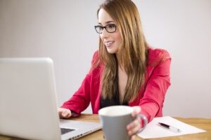 A screenshot of a woman smiling, holding a cup of coffee, and working on a laptop.