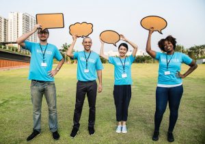 Four young adults in blue t-shirts reading "VOLUNTEER" smiling and holding cardboard cut-outs of speech bubbles over their heads.