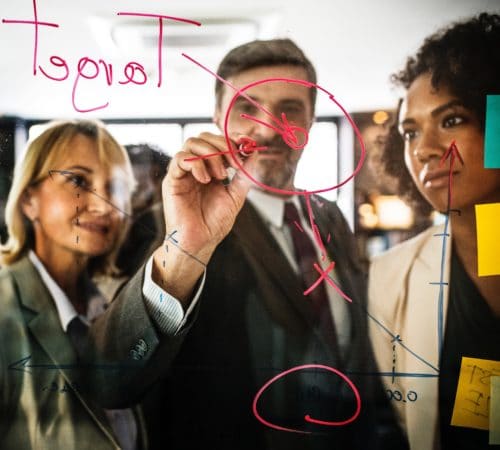 An image from behind a glass screen showing three businesspeople collaborating on a project; one writes on the glass screen with a marker while the other two watch and analyze. Collaborating on a project is a valuable transferable job skill.