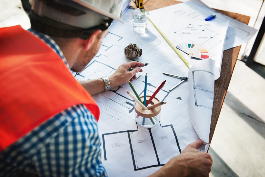 An architect wearing a construction hat and vest bends over a messy table, cluttered with tools, to work on a drawing.