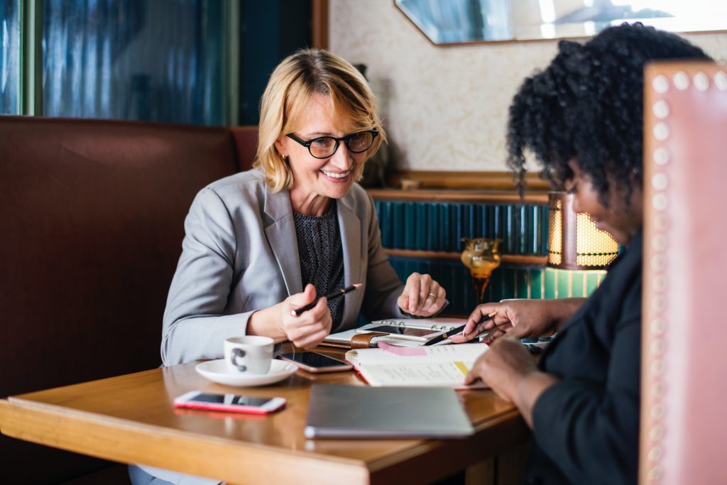 Two women discussing a resume across a table; the woman facing the camera smiles while gesturing to a document (presumably a resume) in the middle of the table.