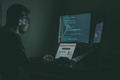 An image of a man in a dark room in front of 3 computer screens filled with coding, demonstrating an example of someone who would use a coding bootcamp resume.