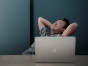 A smiling professionally dressed young man reclining with a look of success in front of a laptop.