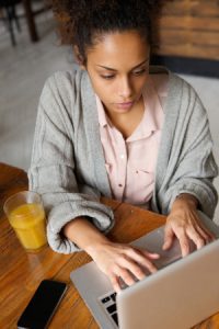 A determined-looking woman sits at a table typing on a laptop
