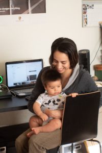 A stock photo of a smiling woman holding a baby in front of her laptop, illustrating the possibility of earning a certification to fill a 2020 Covid resume gap.