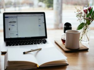 A stock photo of a laptop open to a word processor next to a cup of coffee and an open notebook with a pen, illustrating the importance of writing a cover letter in 2021.