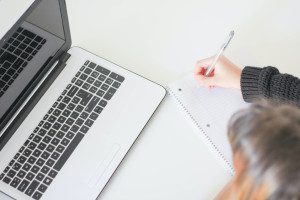 A photo of a laptop with a person writing in an notebook in front of it, demonstrating the value of a using bullet points in a cover letter.