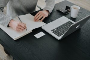 person writing notes in a journal in front of computer